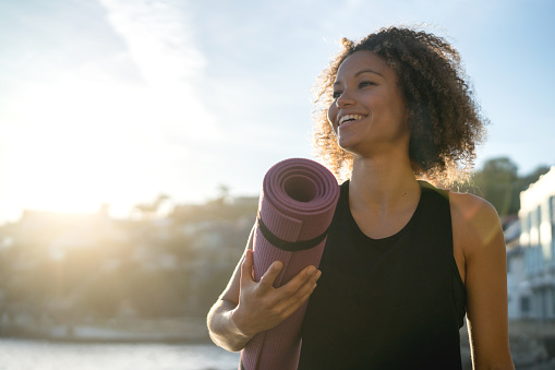 Portrait of a fit woman holding a yoga mat at the beach and looking very happy - wellness concepts