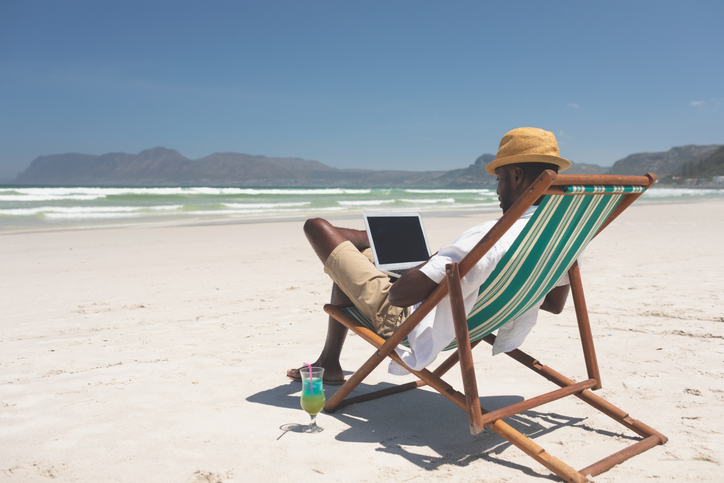 Young man using laptop while sitting at sun lounger at beach - What Men Should Pack For a Caribbean Vacation