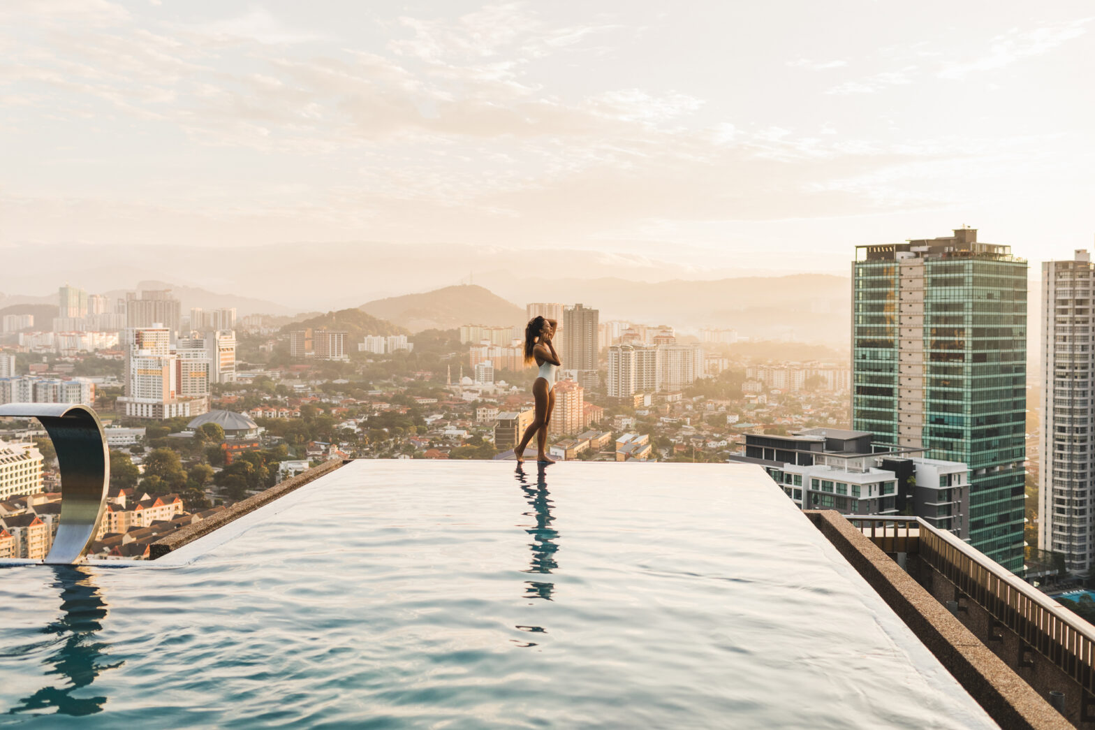 Young woman standing on edge of infinity swimming pool on rooftop view aerial city view at sunset