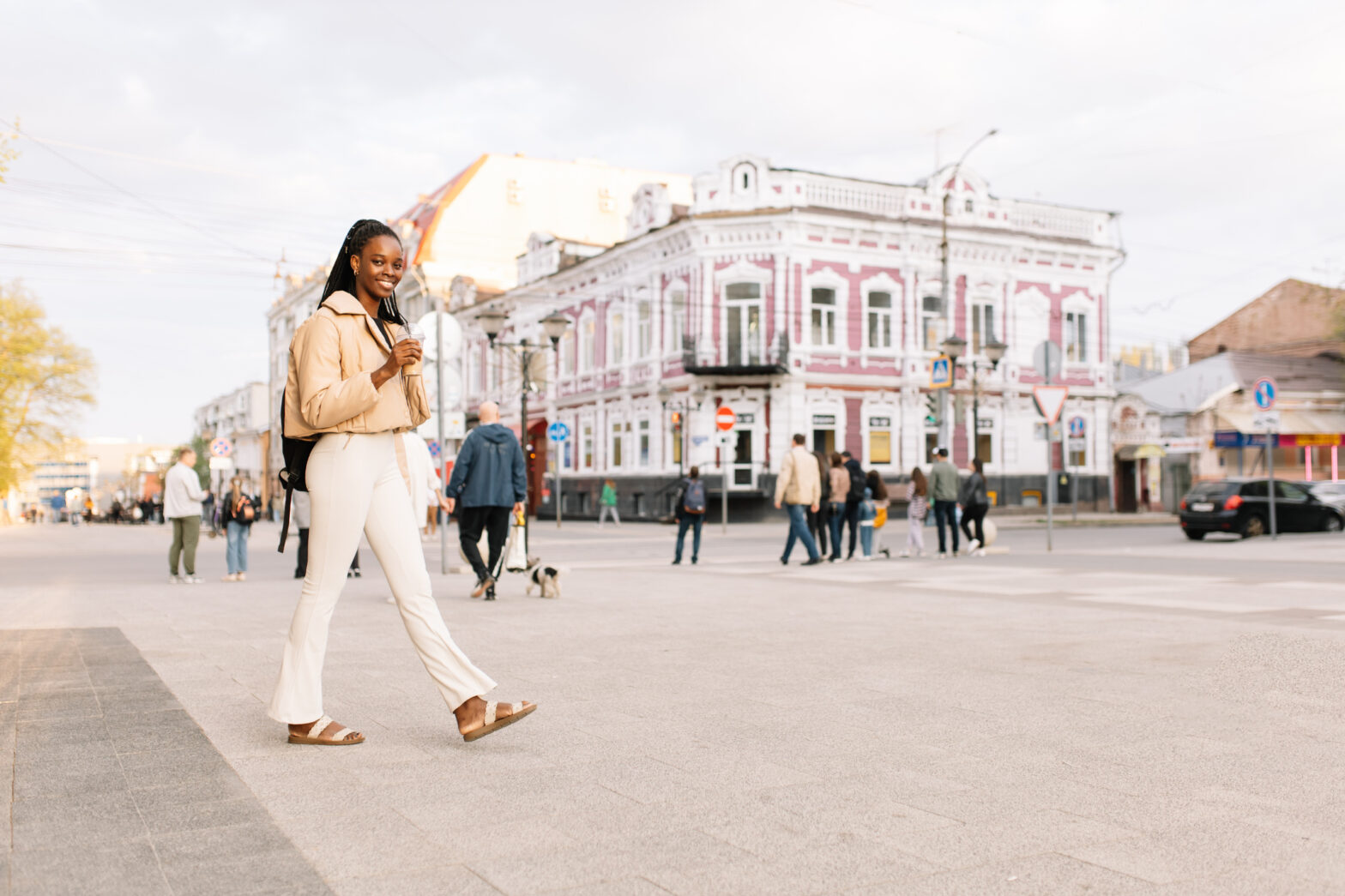 Young black woman with black hair in ponytail in beige jacket and white jeans strolling confidently through town square to see famous clock tower