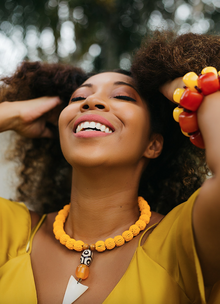 black woman with curly hair smiling and wearing yellow dress with an African jewlery