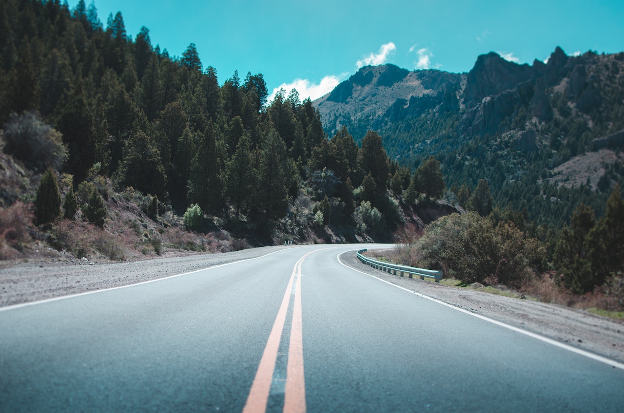 Try to answer these road trip trivia questions with the people you are travelling with to make the drive more enjoyable. Pictured: An open, windy road surrounded by trees and mountains on a sunny day.