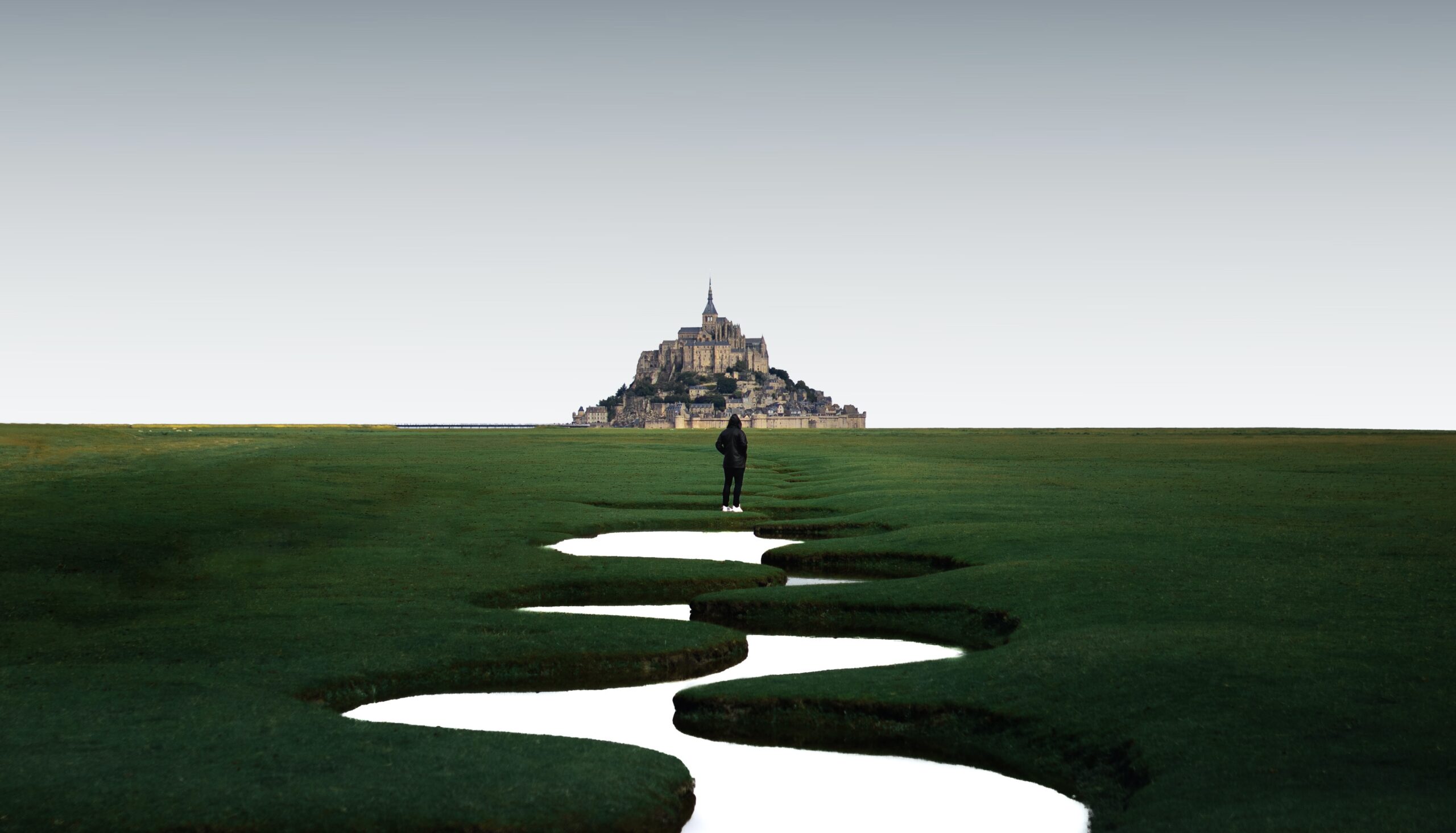 The islet Mont-Saint-Michel is a significant attraction in Normandy. 
Pictured: A long distance photo of the Mont-Saint-Michel from the mainland, with green grass for miles  