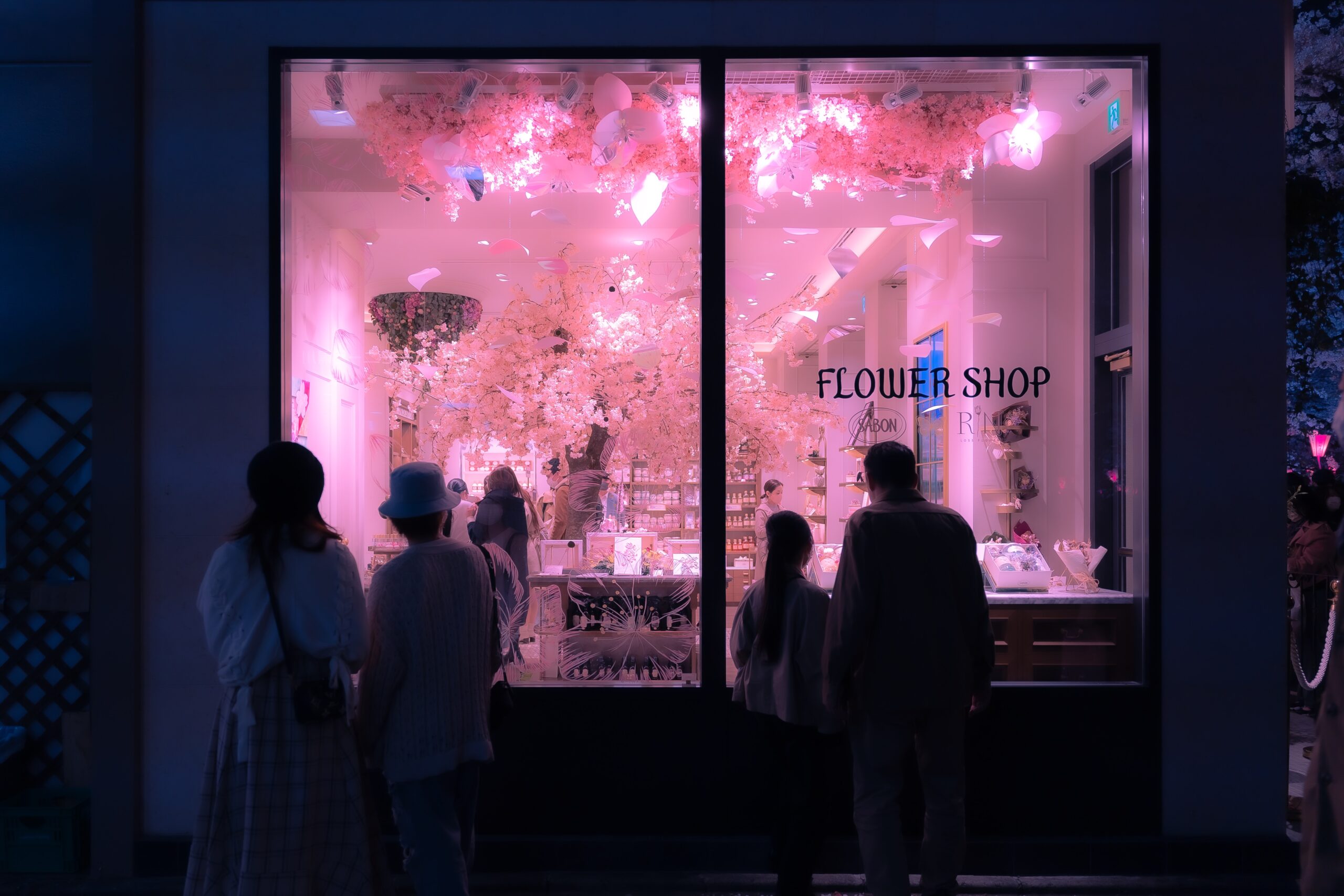 Southern and northern cities of Japan hold sakura in high regard. There are differing times that travelers should visit each region so they can have the best experience.
pictured: A Japanese flower shop with people looking through the shop window that is decorated with pink cherry blossoms 