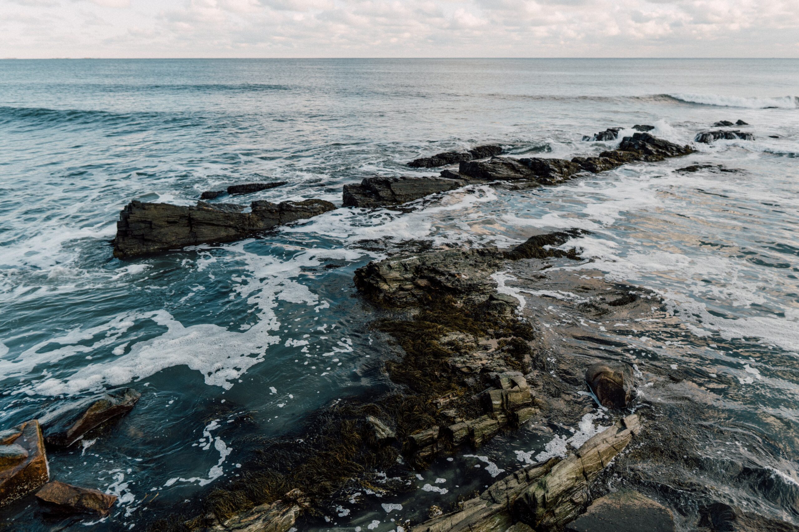 Rhode Island is a seaside state of New England that many travelers should explore on a road trip.
pictured: The warm blue waters of Rhode Island waving over black beach rocks 