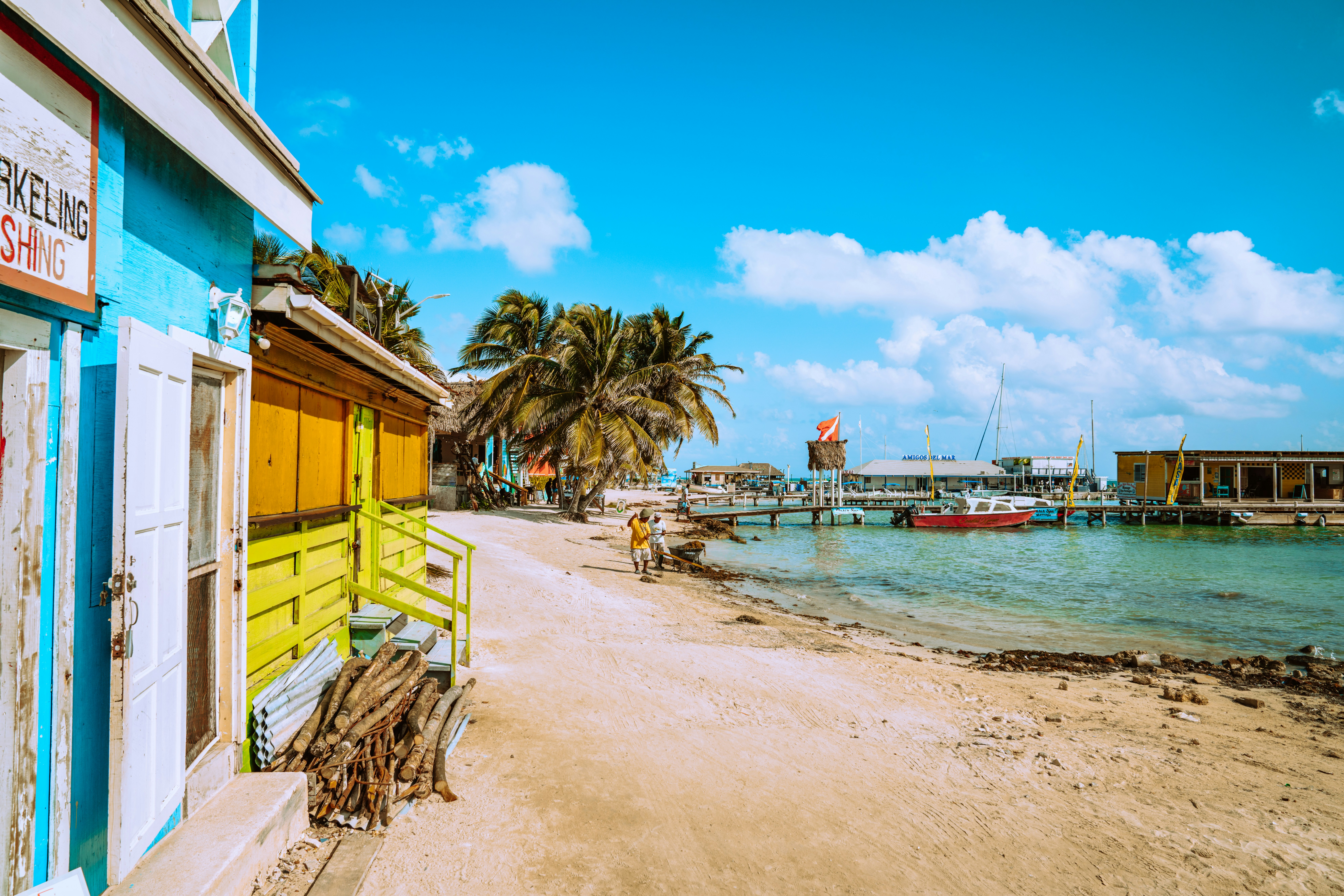 Learn more about the safety level of Belize and ways to avoid danger.
pictured: a white sand beach and turquoise waters near some tourists shops in Belize  