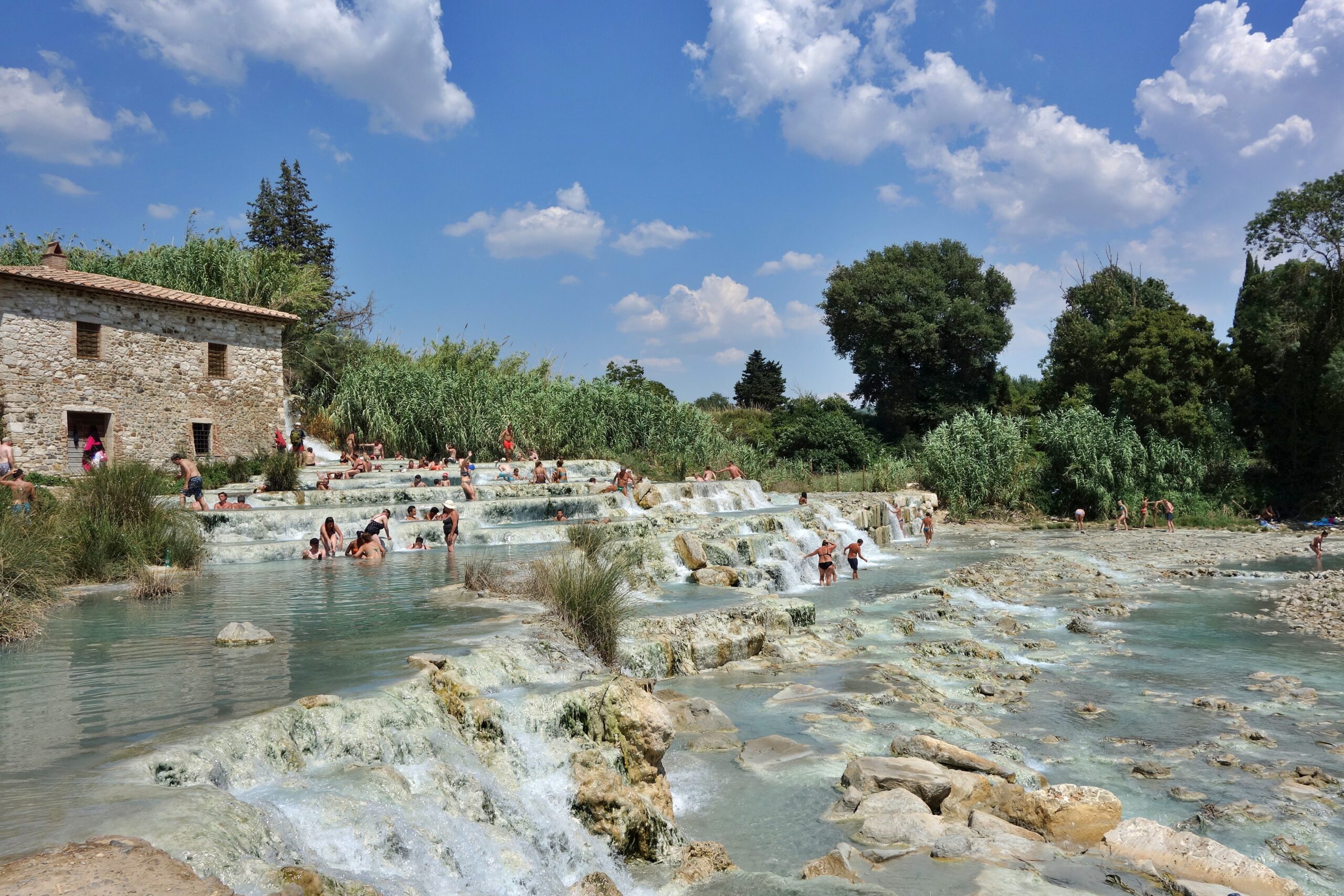 This hot spring in Italy is insanely popular for its serene environment. Check out the benefits that travelers can experience after visiting a hot spring like the one in Saturnia. 
pictured: the Saturnia hot spring with people enjoying the warm waters on a cloudy and sunny day